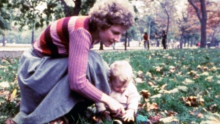 Film still from AS I WAS MOVING AHEAD OCCASIONALLY I SAW BRIEF GLIMPSES OF BEAUTY. A woman and a toddler are sitting in the grass in a park.