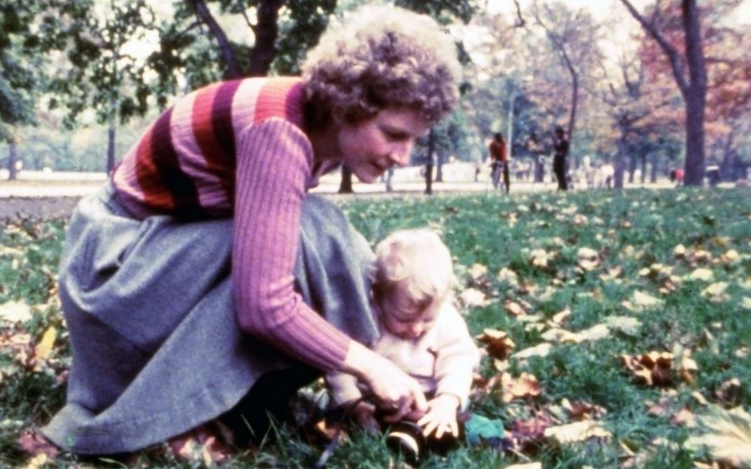 Film still from AS I WAS MOVING AHEAD OCCASIONALLY I SAW BRIEF GLIMPSES OF BEAUTY. A woman and a toddler are sitting in the grass in a park.