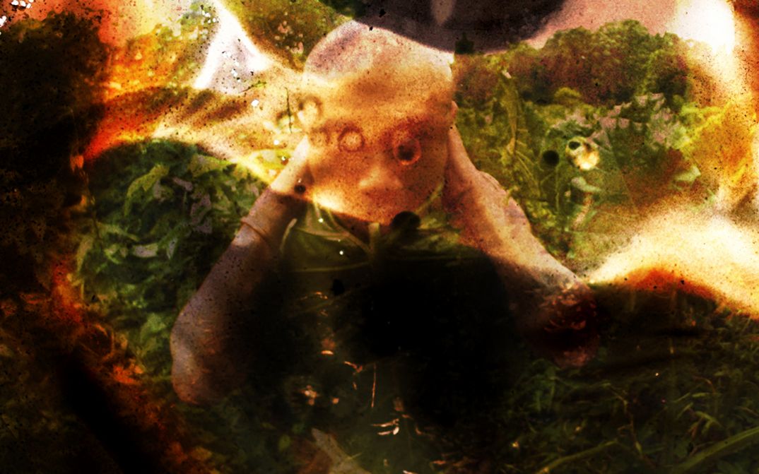 Film Still aus dem Film „Born of the Yam“ von Mark Chua und Lam Li Shuen. Abstraktes, stark verfremdetes Bild einer im Grünen knienden oder kauernden menschlichen Figur mit nach unten geneigtem Kopf, deren Gesicht durch dunkle, körnige Überlagerungen und warme Orange-, Braun- und Grüntöne kaum erkennbar ist.