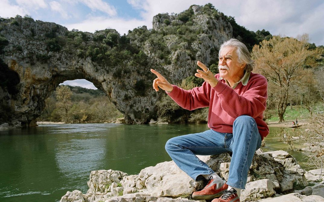 Film still from CAVE OF FORGOTTEN DREAMS: An elderly man sits by a river in front of rock formations with explanatory gestures.