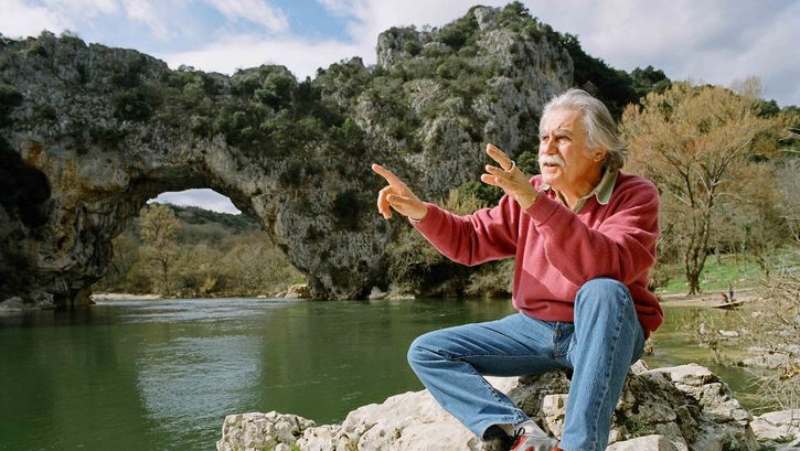 Film still from CAVE OF FORGOTTEN DREAMS: An elderly man sits by a river in front of rock formations with explanatory gestures.