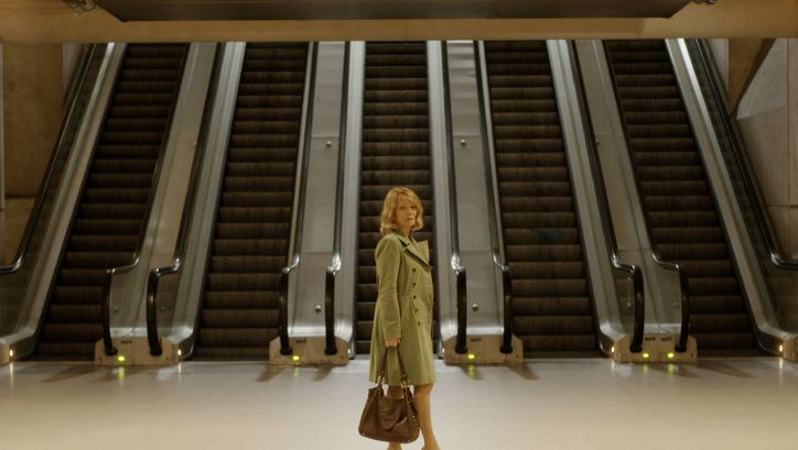 Film still from GARE DU NORD: A woman in a coat and handbag stands in a hall in front of five adjacent escalators.