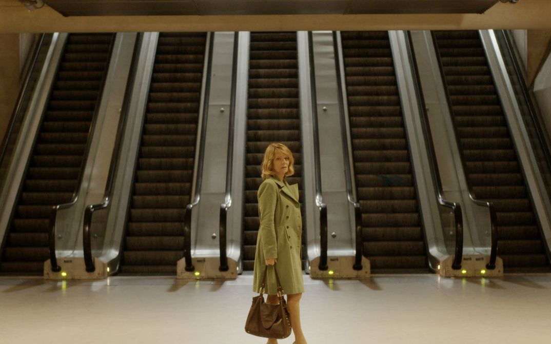 Film still from GARE DU NORD: A woman in a coat and handbag stands in a hall in front of five adjacent escalators.