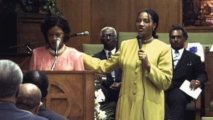 Film still from SUD: Two women are standing at a pulpit with microphones, with other people behind and in front of them.