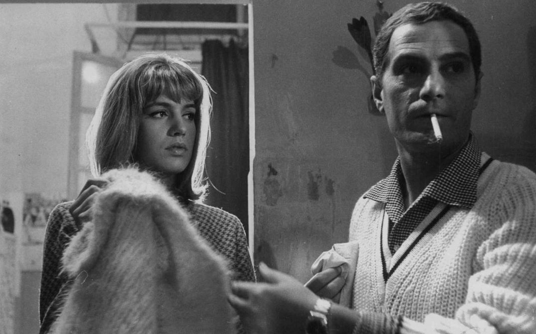 Filmstill aus LA PARMIGIANA: Eine junge Frau und ein Mann in einem Zimmer. Sie schaut ihn an, er ist leicht von ihr abgewandt und hat eine Zigarette im Mund.
