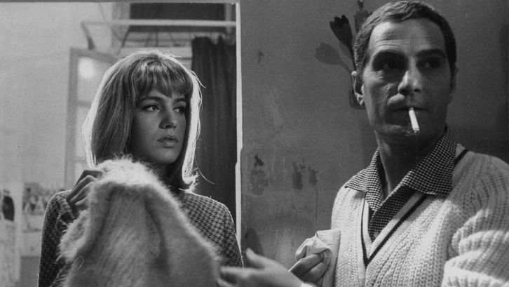 Filmstill aus LA PARMIGIANA: Eine junge Frau und ein Mann in einem Zimmer. Sie schaut ihn an, er ist leicht von ihr abgewandt und hat eine Zigarette im Mund.