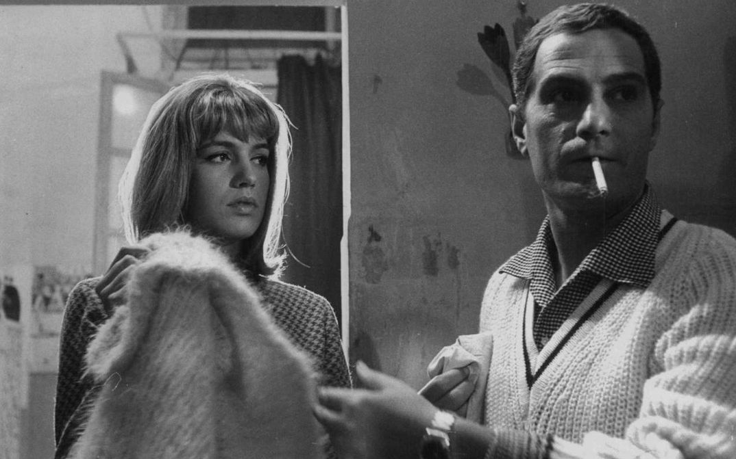 Filmstill aus LA PARMIGIANA: Eine junge Frau und ein Mann in einem Zimmer. Sie schaut ihn an, er ist leicht von ihr abgewandt und hat eine Zigarette im Mund.