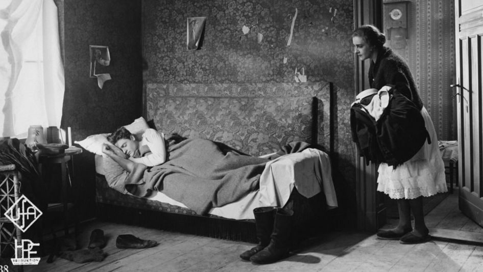 Filmstill aus ZUFLUCHT. Eine Frau blickt in ein Zimmer, in dem ein Mann im Bett schläft.
