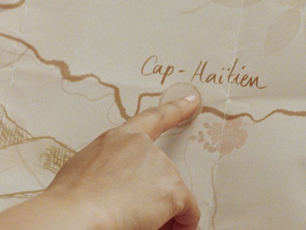 Still from the film "Cette Maison (This House)" by Miryam Charles. A hand points at a close up of a map, with the text "Cap-Haïtian"