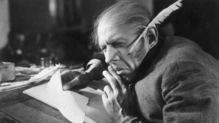 Film still from THE OVERCOAT: An old man sits at a desk, holding a piece of paper in his hand. A quill pen is tucked behind his ear.