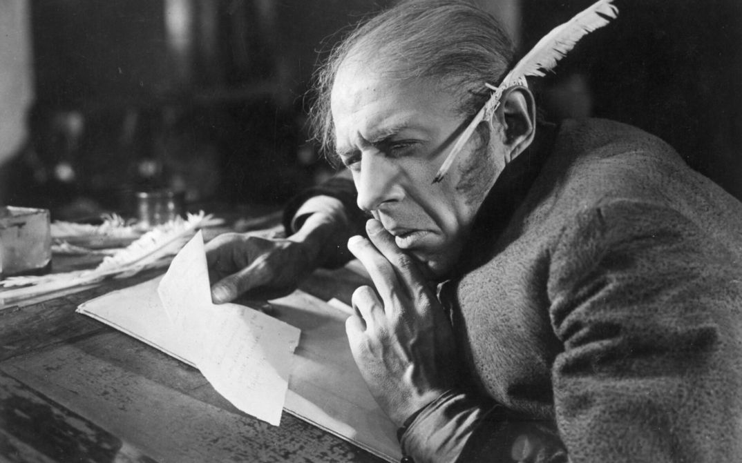 Film still from THE OVERCOAT: An old man sits at a desk, holding a piece of paper in his hand. A quill pen is tucked behind his ear.