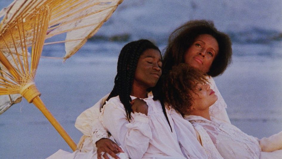 Film still from DAUGHTERS OF THE DUST: Three Black women dressed in summery white clothing are sitting close together by the sea, which is visible in the background, blurred. To their left, a portion of a beach umbrella is visible.