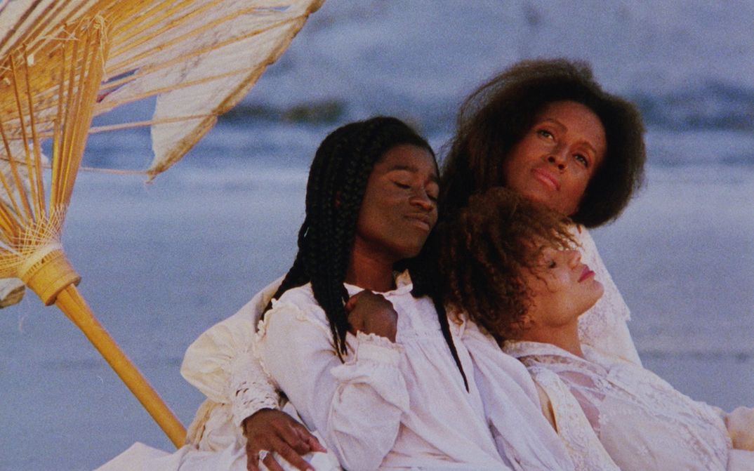 Filmstill aus DAUGHTERS OF THE DUST: Drei Schwarze Frauen in sommerlicher weißer Kleidung sitzen eng zusammen am Meer, das unscharf im Hintergrund zu sehen ist. Links von ihnen befindet sich angeschnitten ein Sonnenschirm.