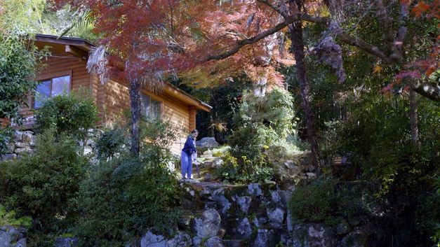 Person steht vor einem Holzhaus in einem herbstlichen Garten mit Bäumen, Felsen und dichtem Grün.