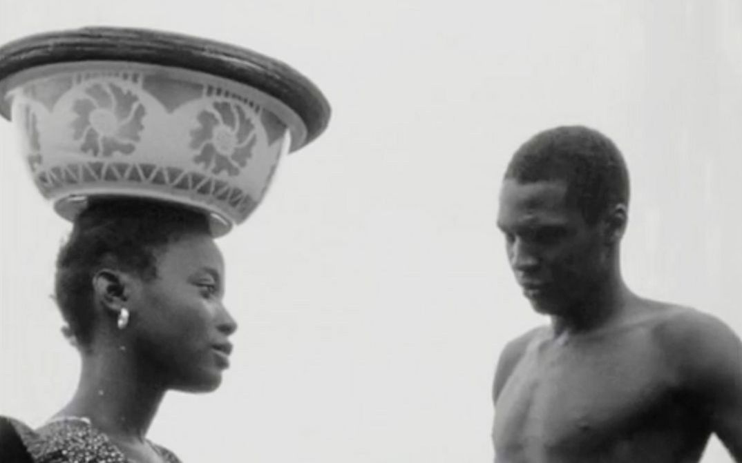 Filmstill aus "Kaddu Beykat" von Safi Faye. zu sehen ist eine Schwarz-Weiß-Aufnahme auf der der Kopf einer Frau zu sehen ist, die eine Schale auf dem Kopf trägt, und ein Mann, der zu ihr gewandt steht.