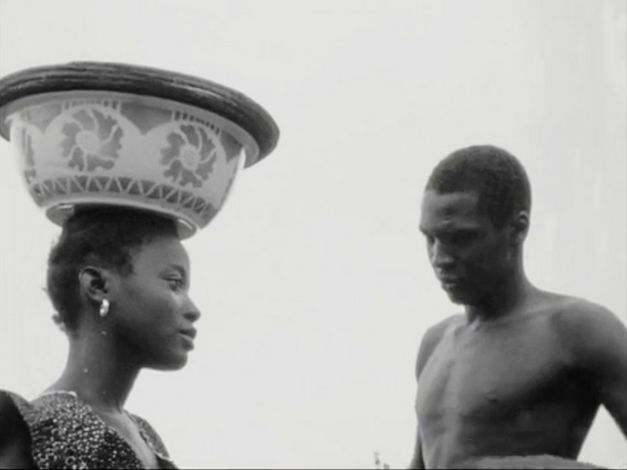 Filmstill aus "Kaddu Beykat" von Safi Faye. zu sehen ist eine Schwarz-Weiß-Aufnahme auf der der Kopf einer Frau zu sehen ist, die eine Schale auf dem Kopf trägt, und ein Mann, der zu ihr gewandt steht.