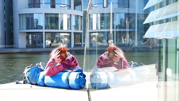 Film still from AUFBRUCH: A young person is sitting by a river, and through a reflection she can be seen twice. There is also a modern building with a glass façade on the other bank of the river. 