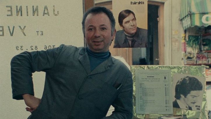 Filmstill aus DAGUERRÉOTYPES: Ein Mann in Arbeitskittel und Hand in die Hüfte gestützt. Hinter ihm Fotos von Männern und ein Gemüsestand.