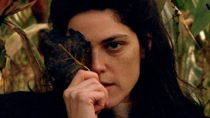 Film still from INVENTION: A young woman with long dark hair holds a leaf in front of her face so that one eye is covered.