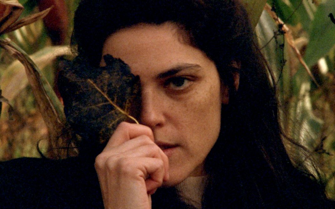Film still from INVENTION: A young woman with long dark hair holds a leaf in front of her face so that one eye is covered.