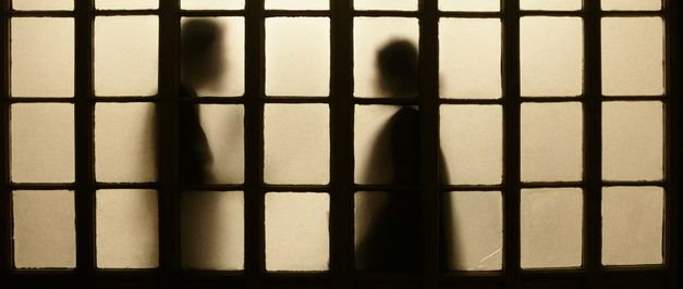 Filmstill aus dem Film "Those Who Do Not Drown" von Naeem Mohaiemen. Man sieht die Silhouetten zweier Menschen in einem erleuchteten Raum durch einen trüben Sichtschutz.