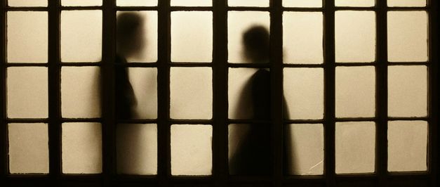 Still from the film "Those Who Do Not Drown" by Naeem Mohaiemen. The silhouettes of two people in a lighted room through a dim screen.