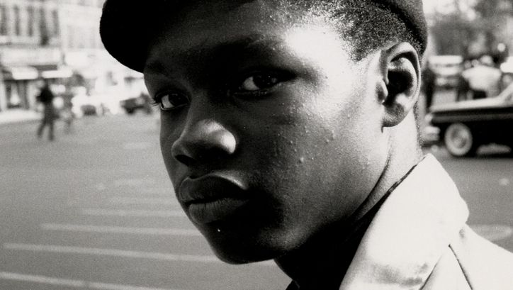 Filmstill aus THE COOL WORLD: Ein Mann mit Schiebermütze von der Seite. Er schaut auf etwas. Im Hintergrund eine Straße, Autos und Passant*innen.