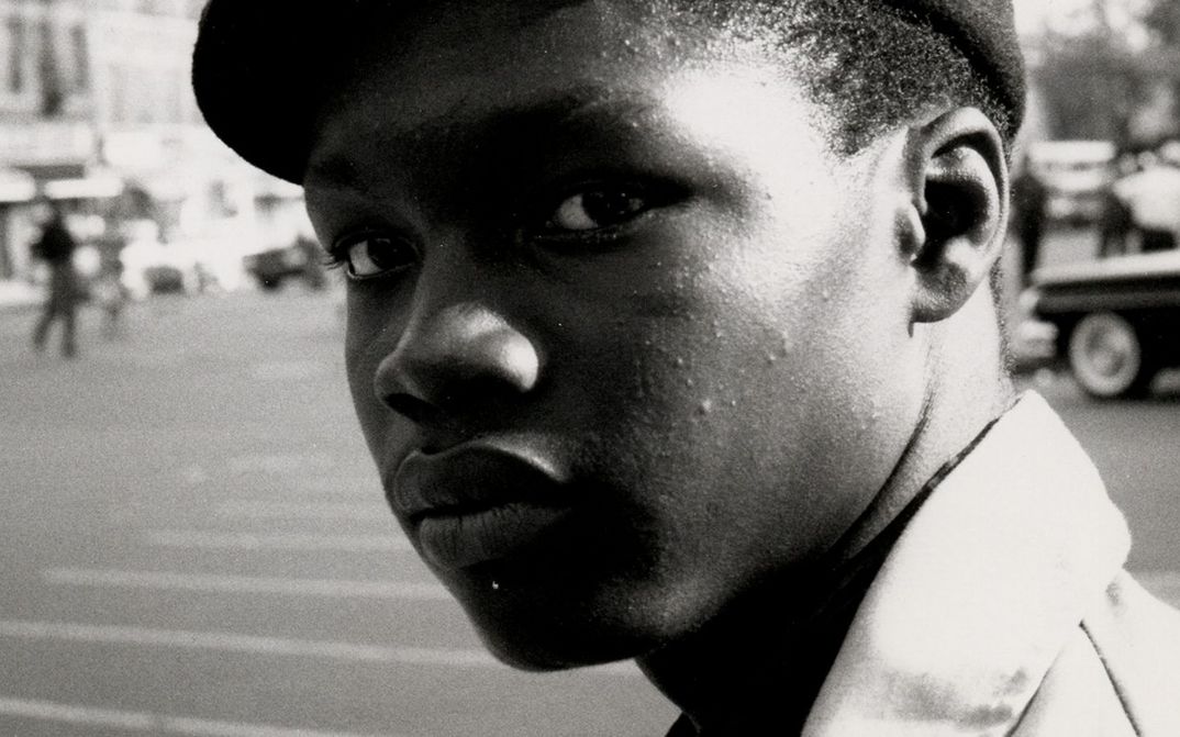 Filmstill aus THE COOL WORLD: Ein Mann mit Schiebermütze von der Seite. Er schaut auf etwas. Im Hintergrund eine Straße, Autos und Passant*innen.