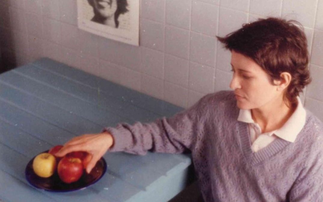 Film still from UM ADEUS PORTUGUÊS: A young woman is sitting at a table. She reaches for the apples on a plate on the table.