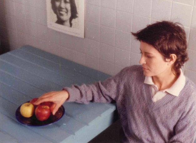Film still from UM ADEUS PORTUGUÊS: A young woman is sitting at a table. She reaches for the apples on a plate on the table.