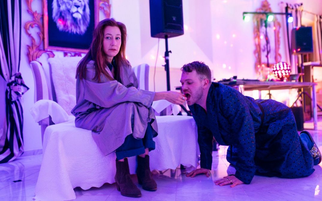 A woman sits in an armchair while a man kneels on all fours and eats from her hand in a colorfully lit room.