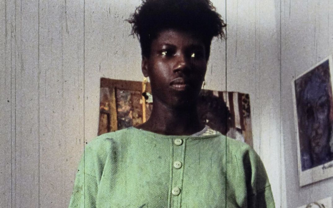 Filmstill aus „A Lover & Killer of Colour“ von Wanjiru Kinyanjui. Nahaufnahme einer Frau. Im Hintergrund hängen Bilder an der Wand.