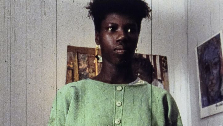 Filmstill aus „A Lover & Killer of Colour“ von Wanjiru Kinyanjui. Nahaufnahme einer Frau. Im Hintergrund hängen Bilder an der Wand.