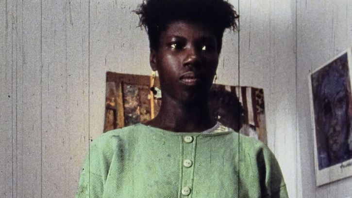 Still from the film „A Lover & Killer of Colour“ by Wanjiru Kinyanjui. Close-up of a woman. In the background, pictures hang on the wall.