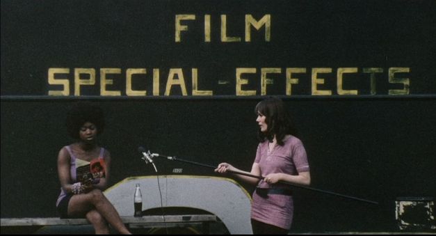 Film still from DARK SPRING. A woman holds a microphone out to another who is reading from a book. In the background the lettering Film Special Effects.