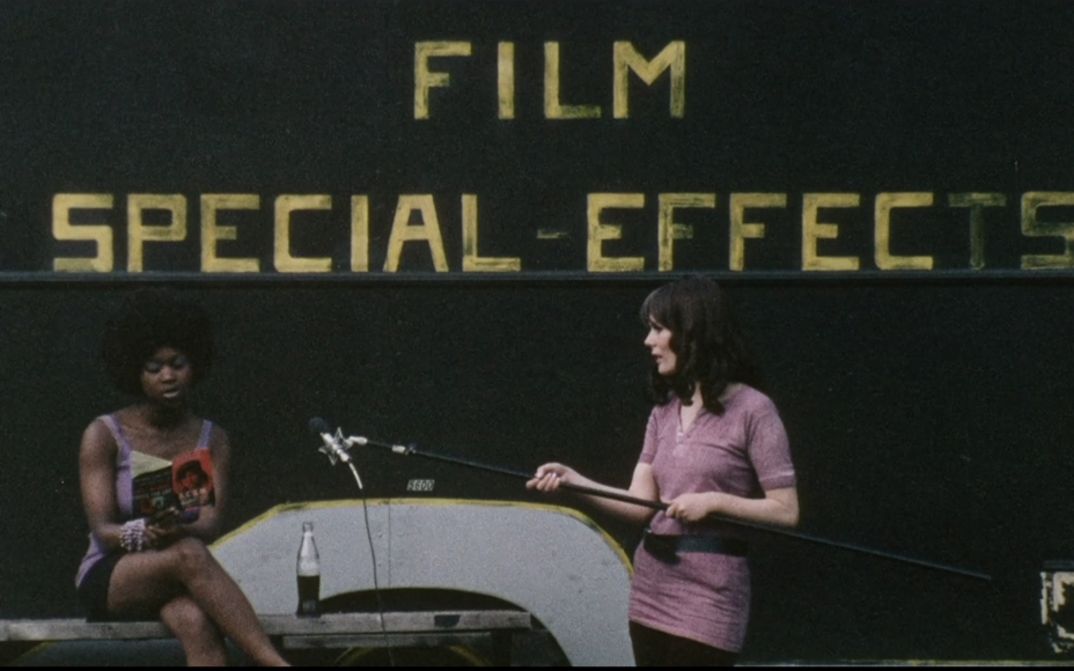 Film still from DARK SPRING. A woman holds a microphone out to another who is reading from a book. In the background the lettering Film Special Effects.