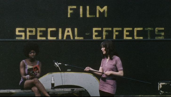 Film still from DARK SPRING. A woman holds a microphone out to another who is reading from a book. In the background the lettering Film Special Effects.