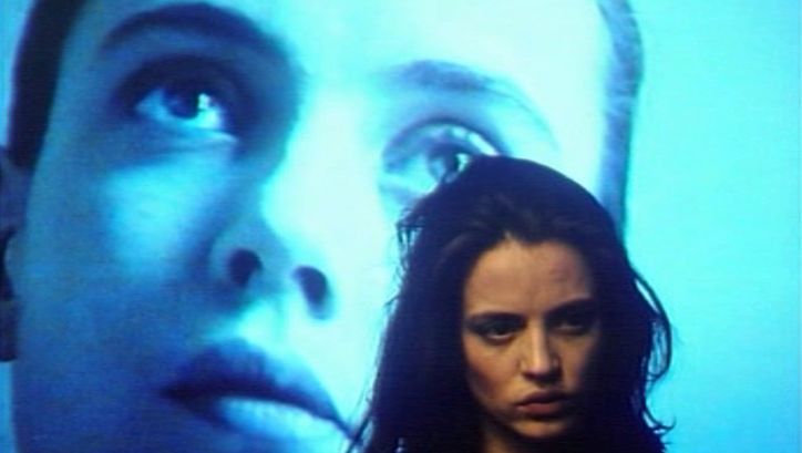 Film still from GINEVRA. Close-up of a woman, in the background a face is projected on a screen.
