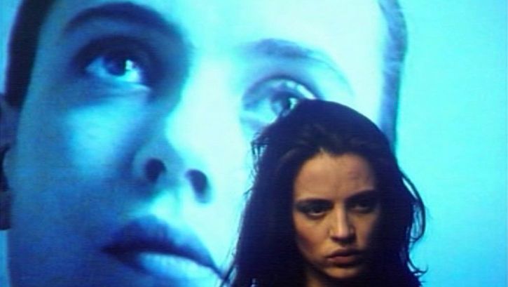 Film still from GINEVRA. Close-up of a woman, in the background a face is projected on a screen.