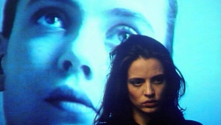 Filmstill aus GINEVRA. Nahaufnahme einer Frau, im Hintergrund ist groß ein Gesicht auf eine Leinwand projiziert.