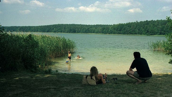 Filmstill from FERIEN: Children in the water of a lake, a man and a woman lying on the meadow in front of it. The sun is shining, the lake is surrounded by reeds and forest.