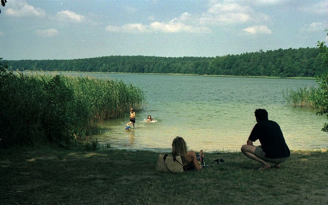 Filmstill from FERIEN: Children in the water of a lake, a man and a woman lying on the meadow in front of it. The sun is shining, the lake is surrounded by reeds and forest.