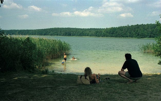 Filmstill aus FERIEN: Kinder im Wasser eines Sees, ein Mann und eine Frau liegen auf der Wiese davor. Die Sonne scheint, um den See ist Schilf und Wald.