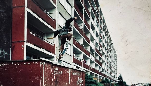 Person springt mit Skateboard vor langem sozialistischen Plattenbau.