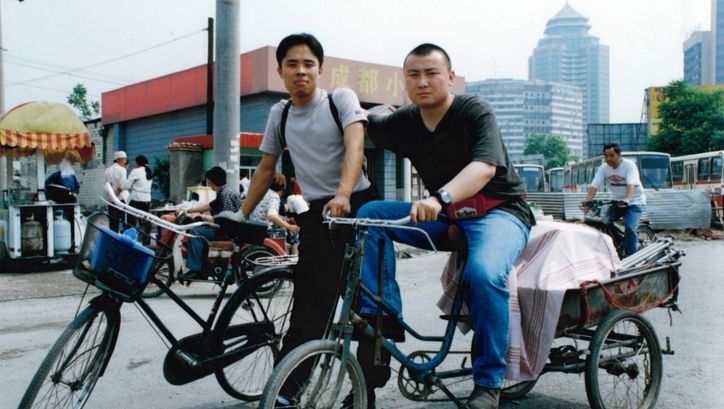 Filmstill aus THERE´S A STRONG WIND IN BEIJING: Zwei junge Männer auf der Straße mit ihren Fahrrädern. Im Hintergrund eine Stadt, Busse und einen Straßenstand.