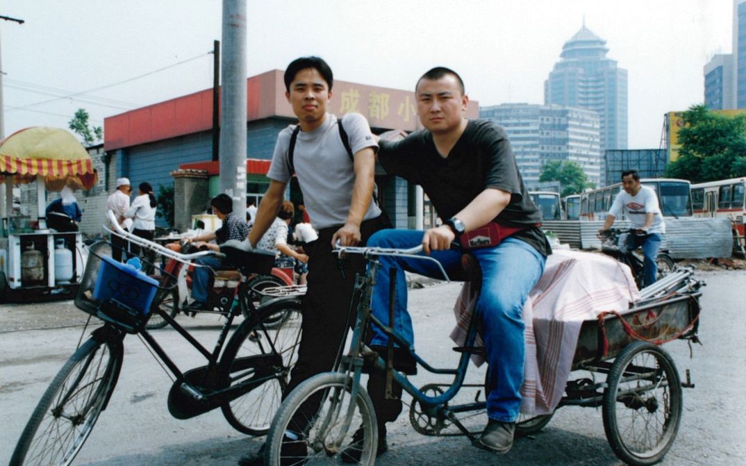Filmstill aus THERE´S A STRONG WIND IN BEIJING: Zwei junge Männer auf der Straße mit ihren Fahrrädern. Im Hintergrund eine Stadt, Busse und einen Straßenstand.