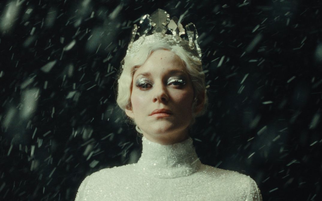 Film still from LA TOUR DE GLACE: A woman dressed as the Snow Queen looks into the camera. She is wearing a white, glittering dress, a white crown, and glittering eye makeup. Snowflakes can be seen around her.