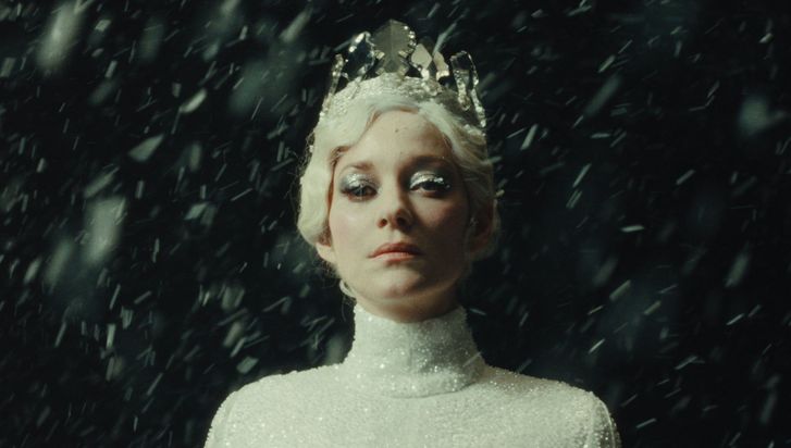 Film still from LA TOUR DE GLACE: A woman dressed as the Snow Queen looks into the camera. She is wearing a white, glittering dress, a white crown, and glittering eye makeup. Snowflakes can be seen around her.