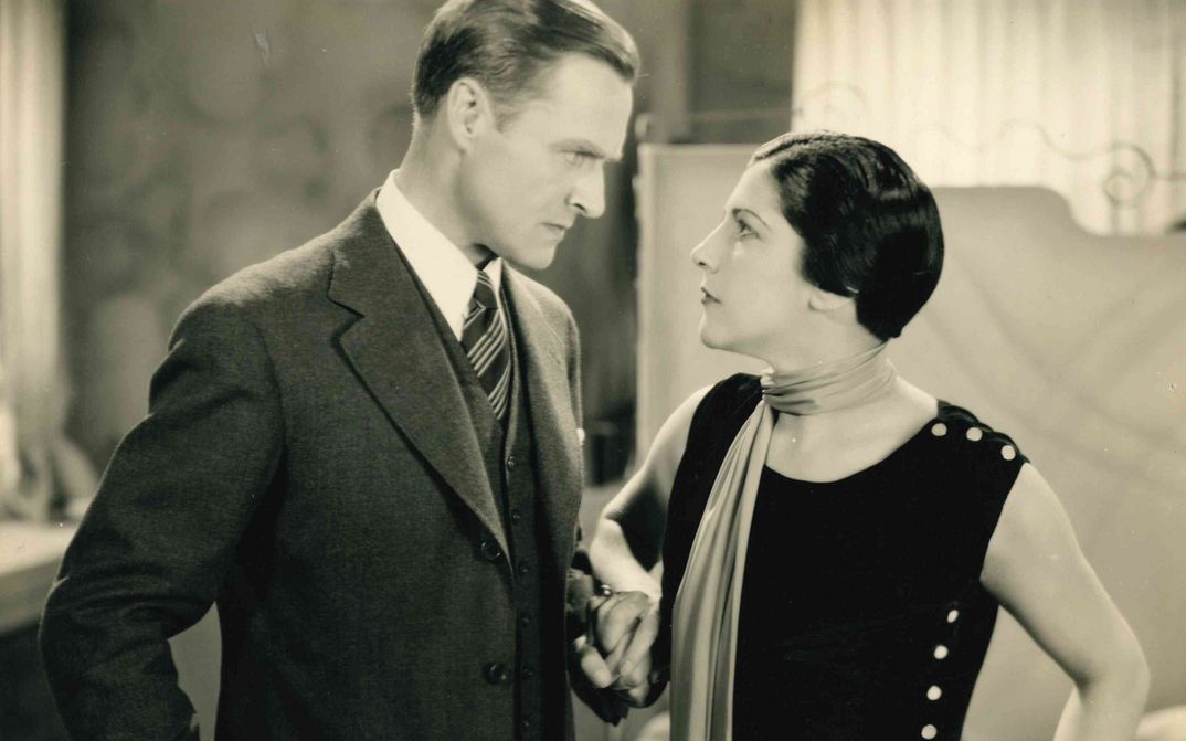 Film still from STRAFSACHE VAN GELDERN: A man and a woman, standing close to each other, look at each other angrily.