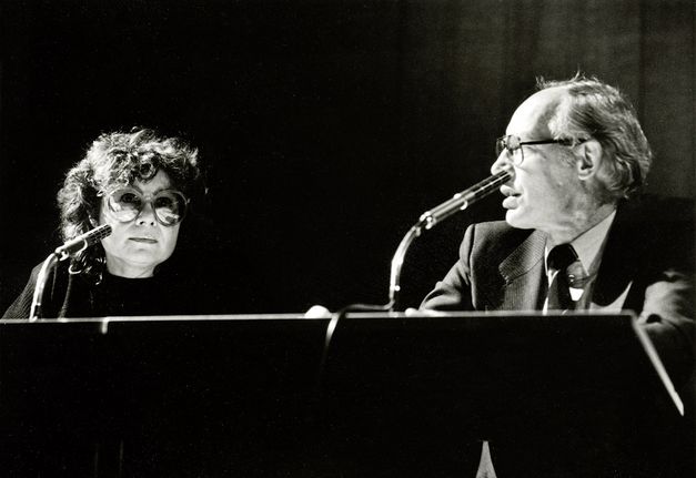 Filmstill aus „Komm mit mir in das Cinema – Die Gregors“ von Alice Agneskirchner. Ein Schwarz-Weiß-Foto einer Frau und eines Mannes auf einem Podium. Vor ihnen stehen Mikros.  